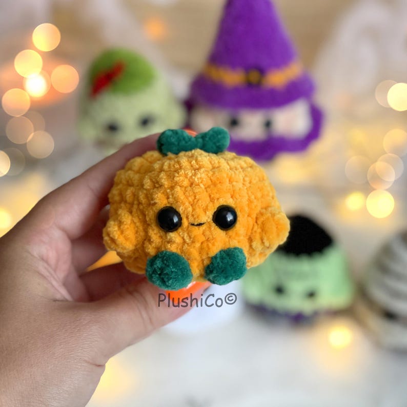 Puede incluir: Una calabaza naranja de peluche con ojos negros y detalles verdes, sostenida en una mano. La calabaza tiene una cara sonriente y un lazo verde. Otros personajes de Halloween de peluche est&aacute;n en el fondo.