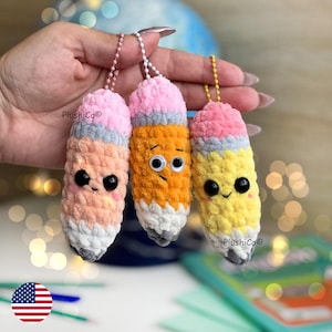 Może przedstawiać: Trzy pluszowe breloczki do kluczy w kształcie ołówków w kolorze brzoskwiniowym, pomarańczowym i żółtym, każdy z różową gumką, szarą opaską i białą końcówką. Ołówki mają ruchome oczy i uśmiechnięte lub neutralne wyrazy twarzy. Każdy breloczek ma metalowy łańcuszek.