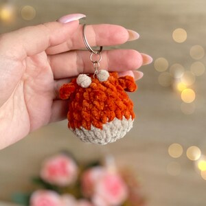 May include: A handmade orange and cream-colored fox keychain. The fox is made of a soft, textured yarn and features two small white pom-poms on top. The keychain is attached to a silver metal ring.