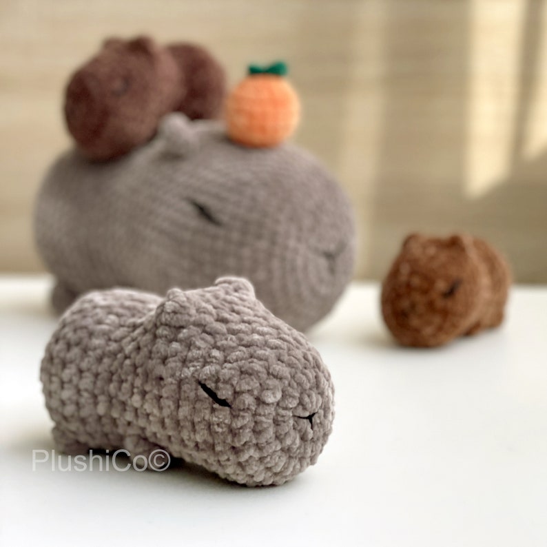 May include: Three crocheted capybara plushies in various sizes. The largest capybara is gray and lying down. A smaller brown capybara is lying down to the right. A small orange pom-pom with a green stem sits on top of the largest capybara.
