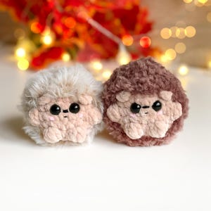 May include: Two handmade crochet hedgehogs. One is light beige with a fluffy exterior, the other is brown. Both have black eyes and a light beige face and paws. The background is blurred with warm colors and bokeh lights.