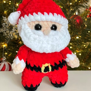 May include: A plush Santa Claus doll made of red, white, black, and beige yarn. The figure features a red hat with a white pom-pom, a thick white beard, black eyes, a black belt with a yellow buckle, and black boots.