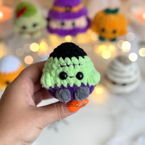 Puede incluir: Un peluche de Frankenstein hecho a mano, de ganchillo, verde, morado y negro, con ojos de bot&oacute;n negros y pies grises. En el fondo se ven otros peluches de ganchillo con tem&aacute;tica de Halloween, como una calabaza, una bruja y una momia.