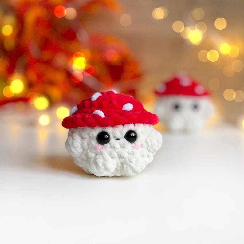 May include: A handmade crochet mushroom toy with a red cap and white spots. The mushroom has a white body, black eyes, and pink cheeks. A second mushroom is visible in the background. The toy is designed to be cute and whimsical.