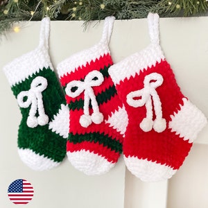May include: Three crocheted Christmas stockings, one green with white stripes, one red with white stripes, and one red with white. Each stocking has a white bow and pom-poms.