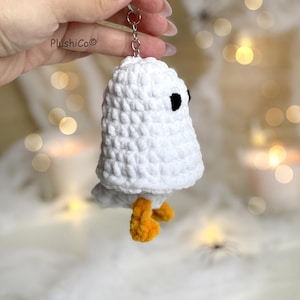 May include: A white crochet ghost keychain with black eyes and orange feet. The handmade ghost is attached to a silver keyring. The PlushiCo brand name is visible in the upper left corner. The background is blurred with soft lighting.