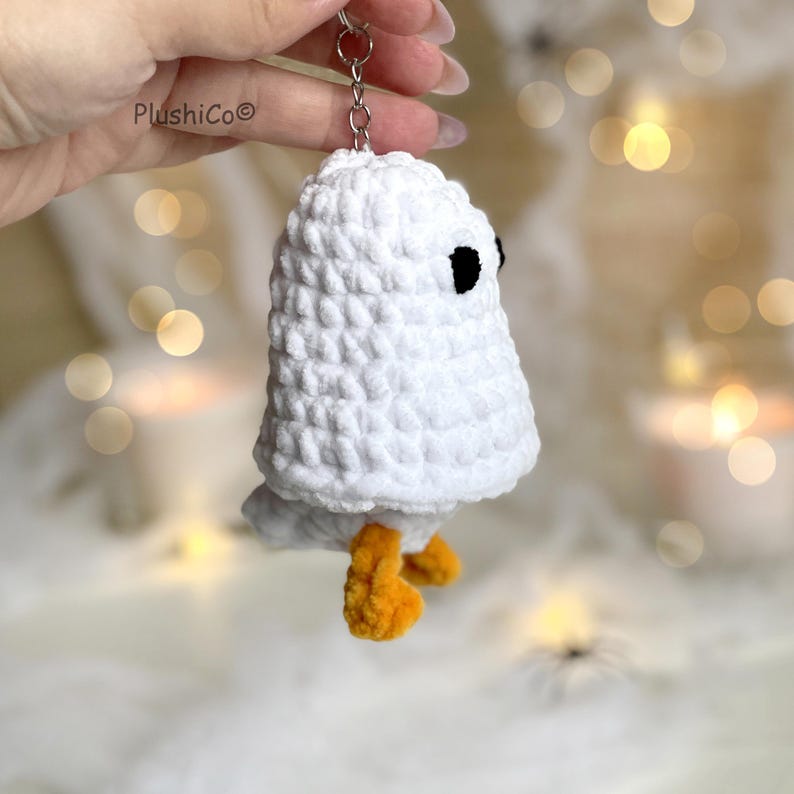 May include: A white crochet ghost keychain with black eyes and orange feet. The handmade ghost is attached to a silver keyring. The PlushiCo brand name is visible in the upper left corner. The background is blurred with soft lighting.
