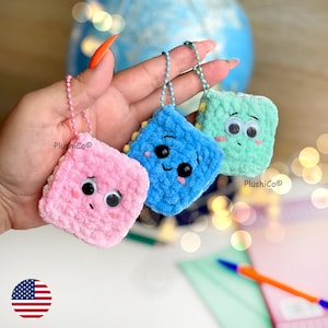 May include: Three crocheted square keychains in pink, blue, and green, each with googly eyes and a smiling or neutral expression. The keychains are attached to silver ball chains. The brand name "PlushiCo" is visible on the keychains.