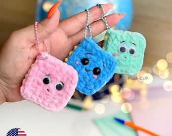 May include: Three crocheted square keychains in pink, blue, and green, each with googly eyes and a smiling or neutral expression. The keychains are attached to silver ball chains. The brand name "PlushiCo" is visible on the keychains.