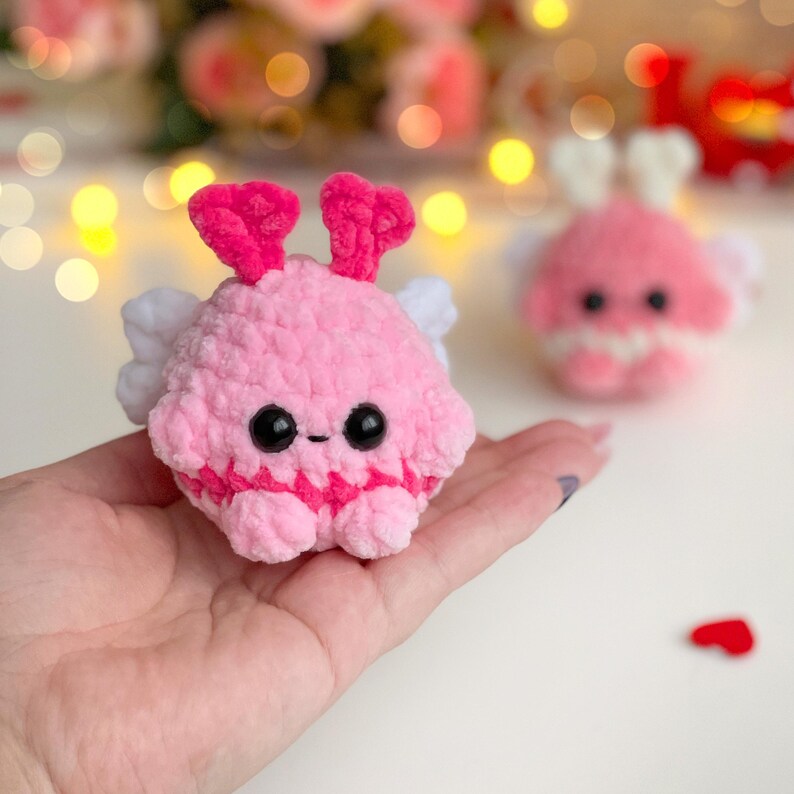 Peut inclure: Une petite cr&eacute;ature ronde crochet&eacute;e en rose avec deux antennes rose vif, des ailes blanches et deux grands yeux de s&eacute;curit&eacute; noirs. Le jouet en peluche pr&eacute;sente une surface en fil textur&eacute; et une petite bouche cousue, pos&eacute; dans une paume.