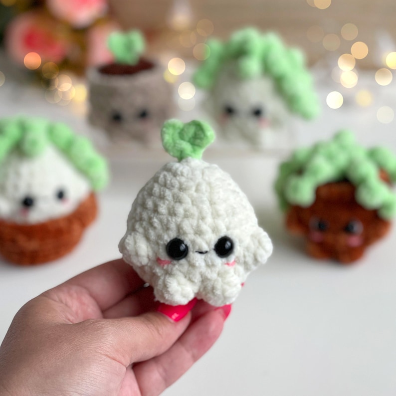 May include: A small, white, crocheted plush toy shaped like a rounded vegetable with a single green leaf on top. It features large black safety eyes and small pink embroidered cheeks. Other similar crocheted plush toys are visible in the background.