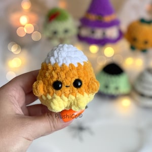 Puede incluir: Un juguete de ganchillo con forma de caramelo de ma&iacute;z sonriente, con ojos negros y una peque&ntilde;a sonrisa. El caramelo de ma&iacute;z es naranja, amarillo y blanco. Otros peluches con tem&aacute;tica de Halloween est&aacute;n en el fondo. El texto "PlushiCo&copy;" es visible.