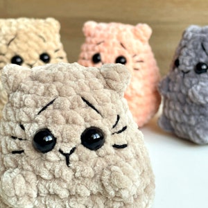 May include: Three crocheted plush toys shaped like cats. The cat on the left is beige, the cat in the middle is peach, and the cat on the right is gray.