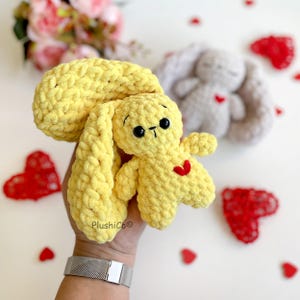 May include: A yellow crocheted bunny with a red heart on its chest. The bunny is being held by a hand with a silver bracelet.