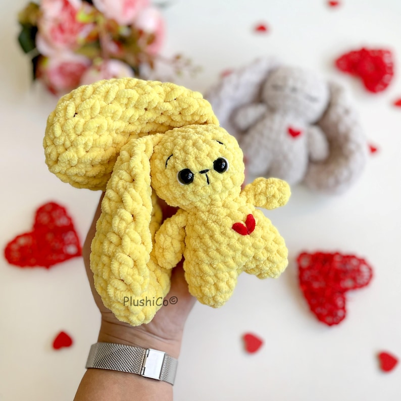 May include: A yellow crocheted bunny with a red heart on its chest. The bunny is being held by a hand with a silver bracelet.