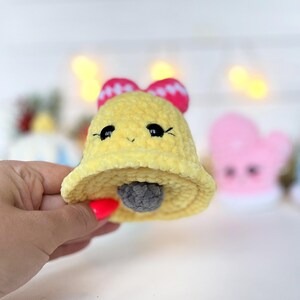 May include: A yellow crocheted bell ornament with a soft, plush texture. The bell features two black plastic eyes, a small embroidered mouth, and a bright pink bow with white polka dots on top. A small gray crocheted ball hangs inside.
