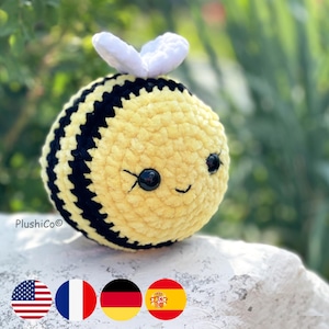 Puede incluir: Un juguete de abeja de peluche tejido a crochet con rayas negras y amarillas, alas blancas y ojos negros de botón. La abeja está sentada en una roca blanca.