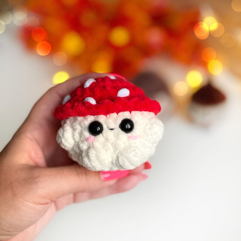 May include: A handmade crochet mushroom with a red cap dotted with white spots and a fluffy white base. The mushroom has large black eyes and pink cheeks, giving it a cute, friendly appearance. The background is blurred with warm, autumnal colors.