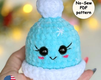 May include: A blue and white crocheted hat with a pom-pom on top. The hat has a cute face with black eyes, pink cheeks, and a white snowflake on the front. The hat is a no-sew PDF pattern.