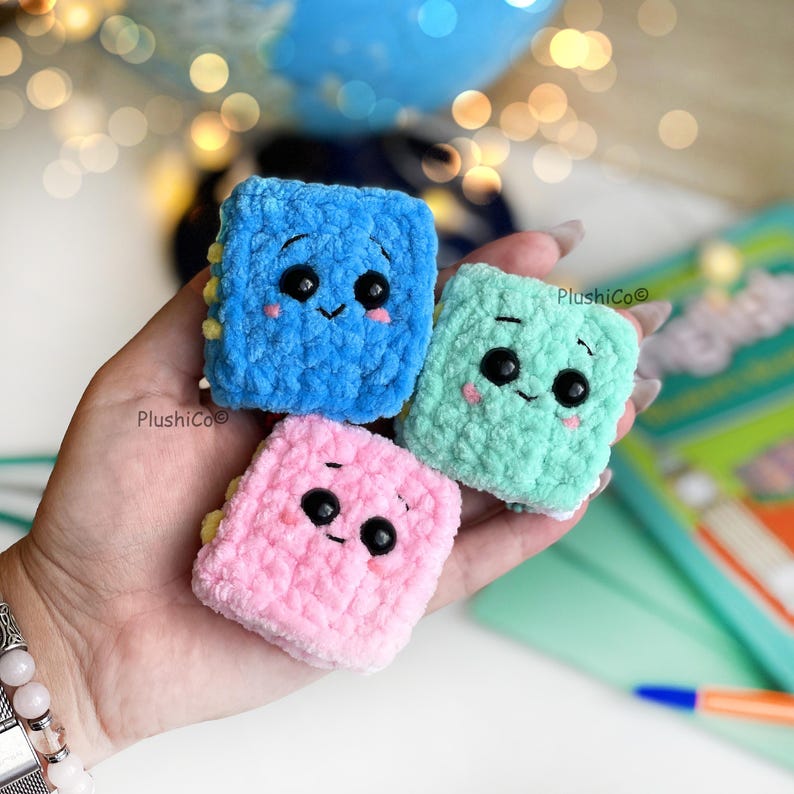 May include: Three small, square, crocheted plush toys in blue, mint green, and pink. Each has a smiling face with black eyes and rosy cheeks. The toys are handmade and have a soft, textured appearance.