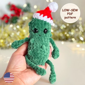 May include: A green crocheted amigurumi toy shaped like a pickle wearing a red and white crocheted Santa hat. The toy has black eyes and a smiling mouth. The text "LOW-SEW PDF pattern" is in the upper right corner of the image.