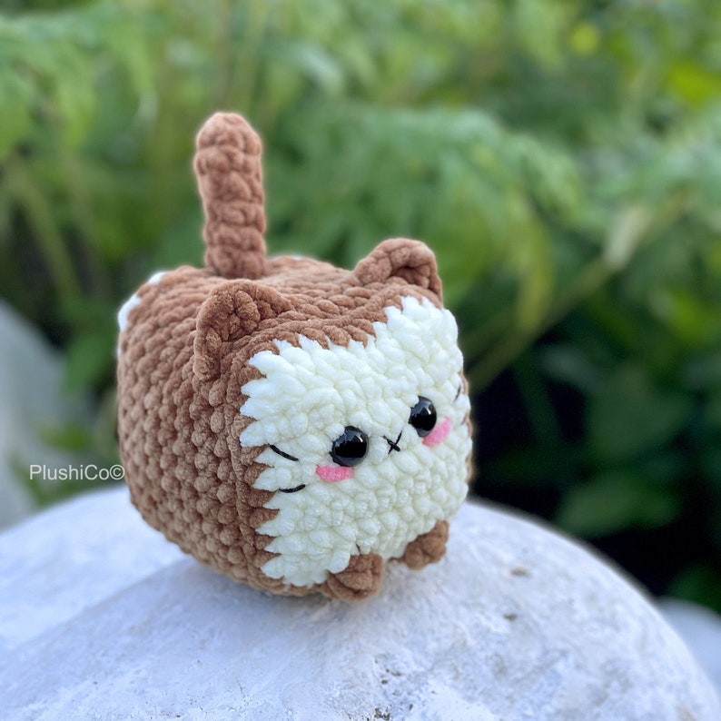 May include: A brown and white crocheted cat plush toy with a cute face and big eyes. The toy has a brown tail and is sitting on a white rock.