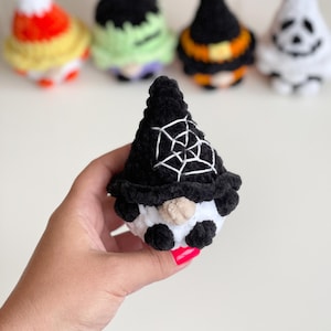 May include: A small, handcrafted crochet gnome featuring a black pointed hat with a white embroidered spiderweb design. The gnome has a round beige nose, a white beard, and small black feet, with other similar crochet figures blurred in the background.