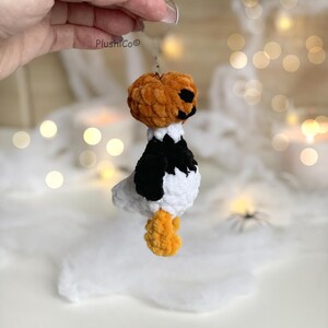 May include: A handmade Halloween-themed plushie featuring a pumpkin head with black facial features, a white body with black accents, and orange feet. The plushie is suspended by a metal loop. The brand name "PlushiCo" is visible.