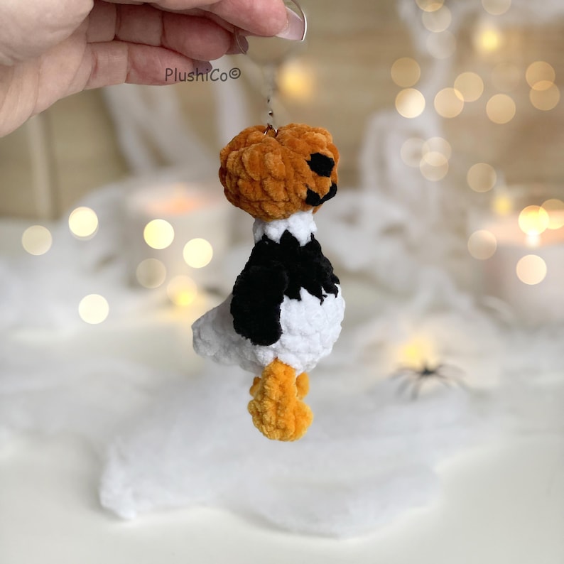 May include: A handmade Halloween-themed plushie featuring a pumpkin head with black facial features, a white body with black accents, and orange feet. The plushie is suspended by a metal loop. The brand name "PlushiCo" is visible.