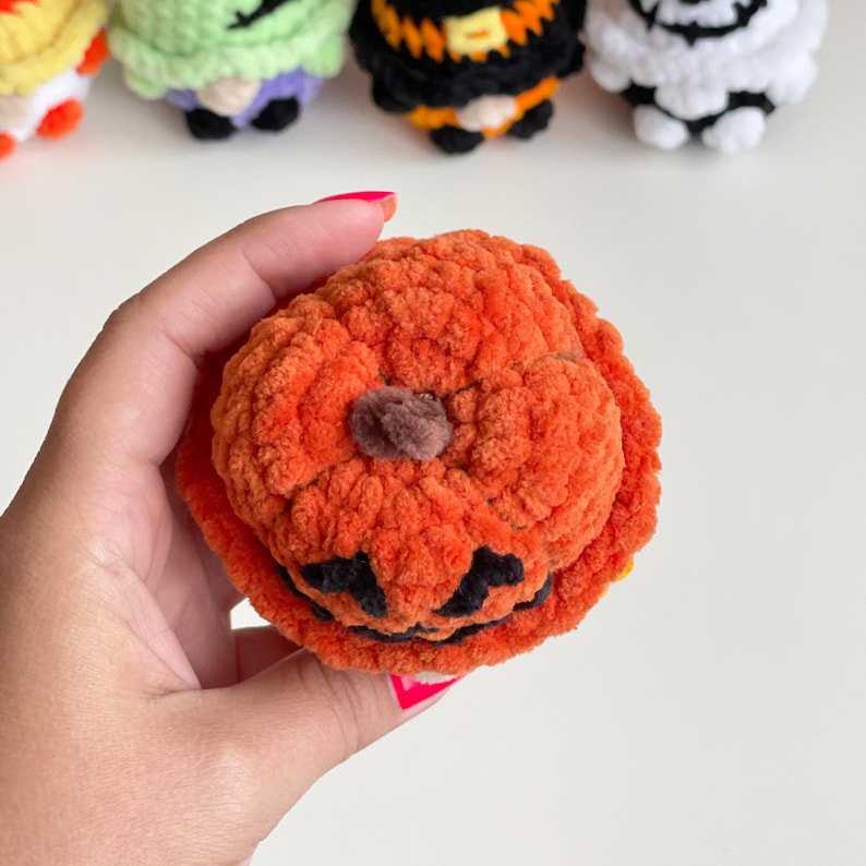 May include: A small, round, orange crocheted pumpkin figure with a textured yarn surface, a brown yarn stem at the top, and a black yarn stitched face featuring a wide, jagged grin and triangular eyes, crafted from soft plush material.