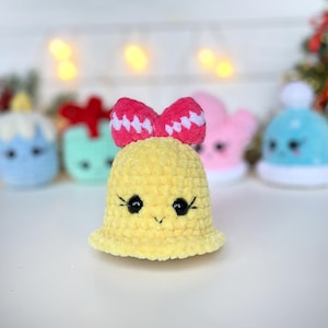 May include: A yellow crochet bell plush toy featuring a bright pink bow with white accents on top. The bell has two black plastic safety eyes with stitched eyelashes and a small stitched mouth, creating a friendly face on the textured yarn surface.