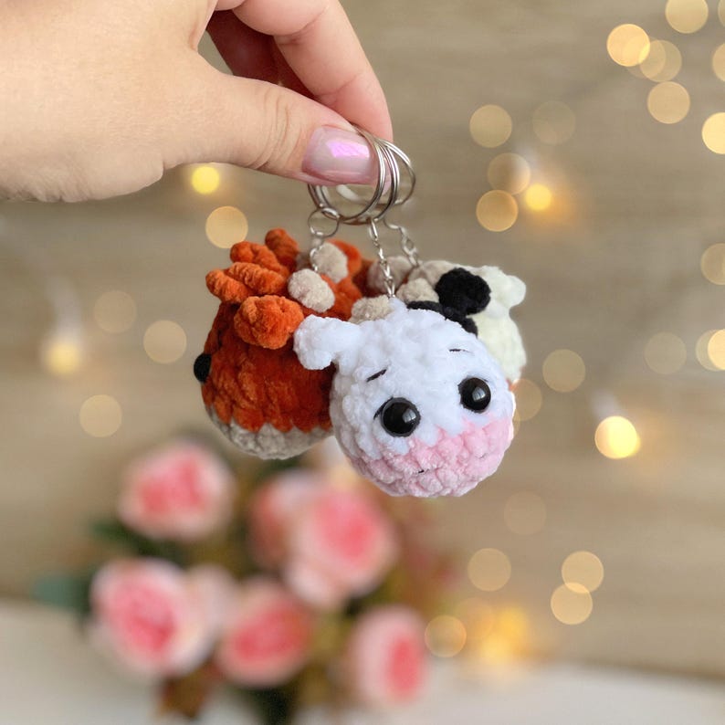May include: A handmade keychain featuring three plush animal figures: a fox, a cow, and a sheep. The fox is orange and white, the cow is white with pink cheeks, and the sheep is white with black spots. The keychains are attached to a silver ring.