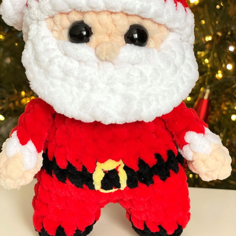 May include: A plush Santa Claus doll crafted from soft red, white, black, and beige yarn. The figure features a white beard, a red hat with a white trim, a black belt with a yellow buckle, and large black plastic eyes. The texture is fuzzy.