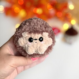 May include: A handmade, plush hedgehog toy with a light brown face and dark brown fur. The hedgehog has black eyes and a small, stitched mouth. The toy is round and soft, perfect for children or as a decorative item.