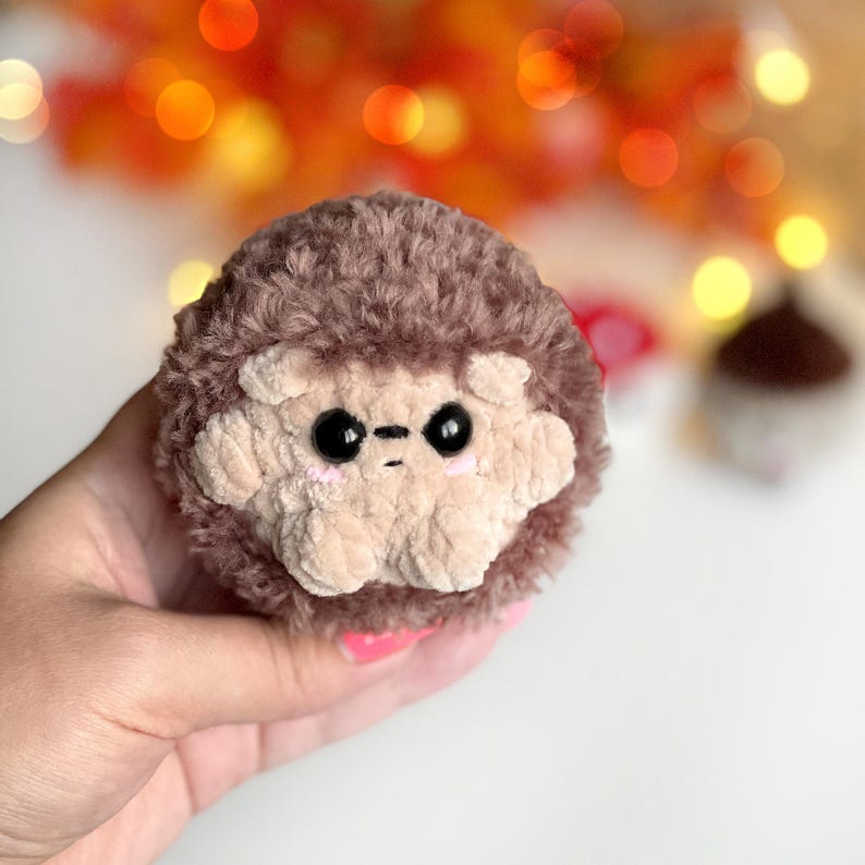 May include: A handmade, plush hedgehog toy with a light brown face and dark brown fur. The hedgehog has black eyes and a small, stitched mouth. The toy is round and soft, perfect for children or as a decorative item.