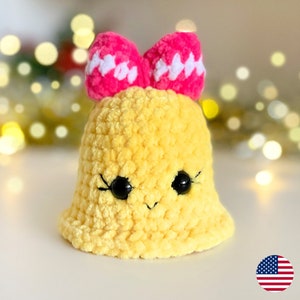 May include: A yellow crocheted bell with a pink bow and black eyes. The bell has a smiling expression.