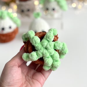 May include: A small, round, crocheted plush toy shaped like a potted plant. The base is a textured, dark brown yarn, while the top features multiple, curly, light green yarn tendrils extending outward, mimicking the appearance of a succulent plant.