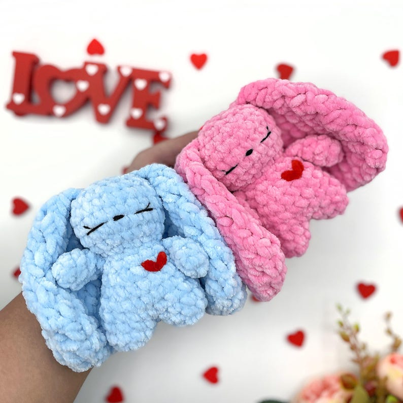 May include: Two crocheted bunny plushies with red hearts on their chests. The bunny on the left is light blue and the bunny on the right is pink.