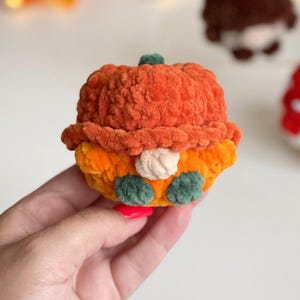 May include: A small, round, plush crochet figure shaped like a pumpkin. The top is a textured orange pumpkin cap with a small green stem. The body is orange with a beige nose in the center and two dark green feet at the bottom, made of soft yarn.
