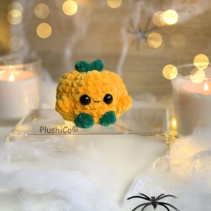 May include: A plush, orange pumpkin with a smiling face, green stem, and green feet. The pumpkin is sitting on a clear surface with a blurred background of candles and spiderweb decorations. The text "PlushiCo" is visible.