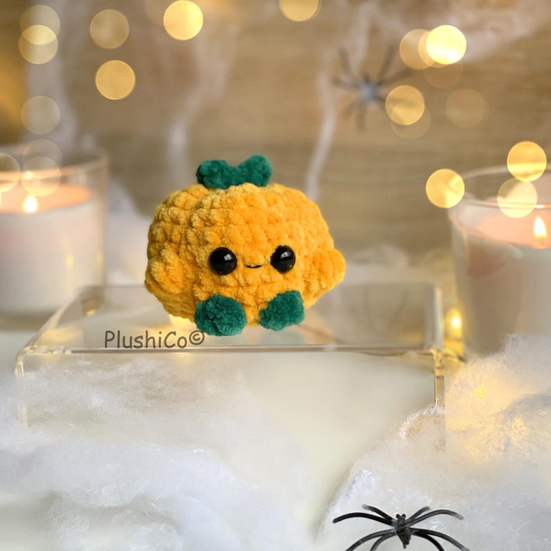 May include: A plush, orange pumpkin with a smiling face, green stem, and green feet. The pumpkin is sitting on a clear surface with a blurred background of candles and spiderweb decorations. The text "PlushiCo" is visible.