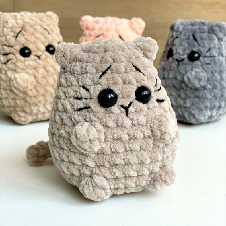 May include: Three crocheted plush cat toys. The cat on the left is beige, the cat in the center is light brown, and the cat on the right is gray. All three cats have black eyes and black embroidered whiskers.