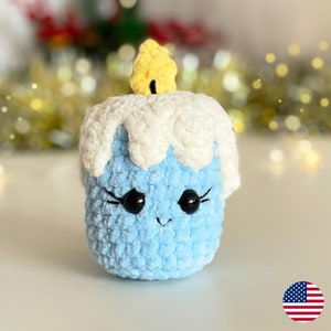 May include: A blue crocheted candle with a white crocheted top and a yellow crocheted flame. The candle has a smiling face with black eyes and eyelashes.
