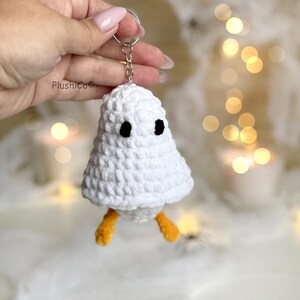 May include: A white crocheted ghost keychain with black eyes and orange feet. The ghost is held by a silver keyring. The keychain is handmade and has a soft, textured appearance. The background is blurred with bokeh lights.