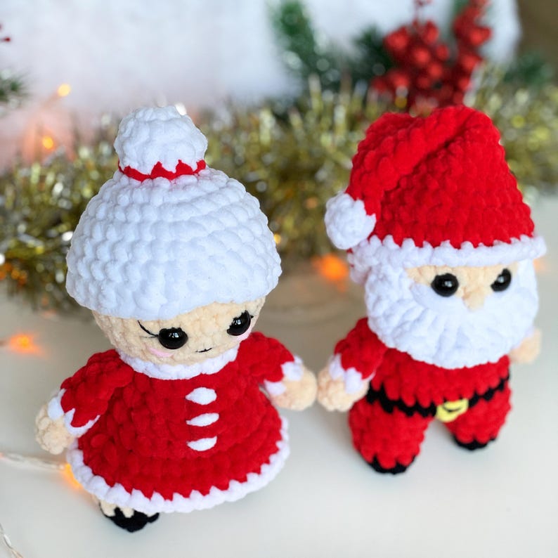 Puede incluir: Un conjunto de mu&ntilde;ecas de crochet de Pap&aacute; Noel y la se&ntilde;ora Claus. Las mu&ntilde;ecas est&aacute;n vestidas de rojo y blanco con ribetes blancos. Pap&aacute; Noel tiene una barba blanca y un gorro rojo. La se&ntilde;ora Claus tiene un gorro blanco y un vestido rojo.