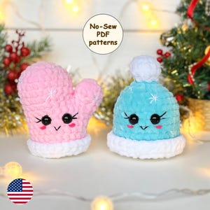 May include: Two crocheted plush toys, a pink mitten and a blue hat, with white trim and snowflake details. Both toys have cute cartoon faces with black eyes and a smiling mouth. The toys are on a white surface with a string of lights in the background. No-Sew PDF patterns.