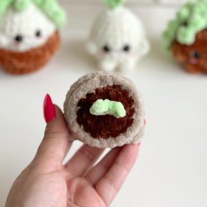 May include: A round, crocheted plush toy featuring a light beige outer ring, a dark brown textured center, and a small light green sprout on top. The plush is crafted from soft yarn and has a circular shape, with other similar plush toys visible in the background.