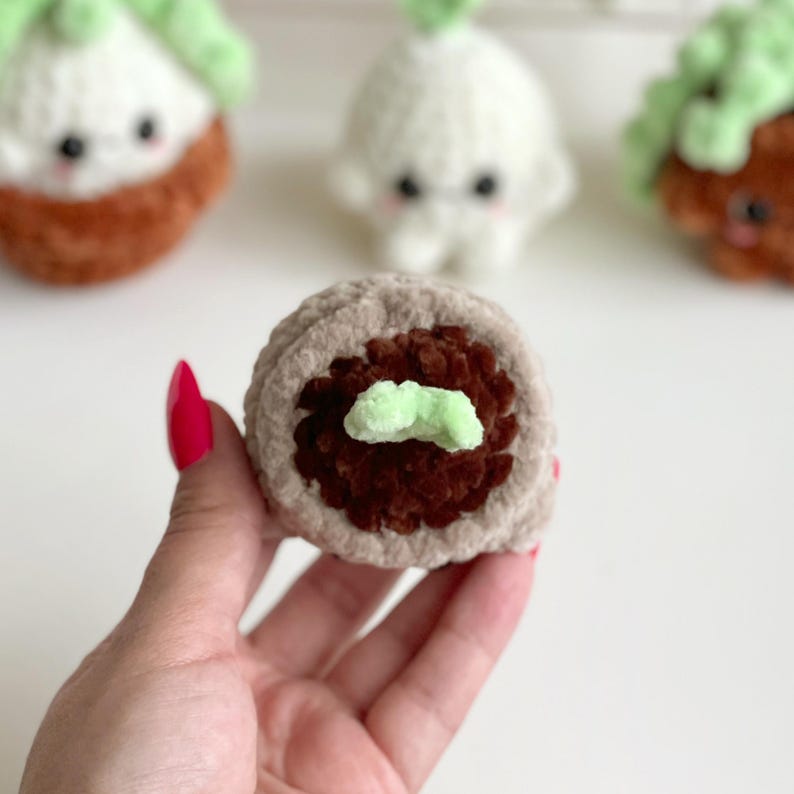 May include: A round, crocheted plush toy featuring a light beige outer ring, a dark brown textured center, and a small light green sprout on top. The plush is crafted from soft yarn and has a circular shape, with other similar plush toys visible in the background.