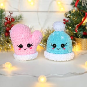 May include: A plush crochet mitten in light pink and a plush crochet beanie in light blue, both featuring white cuffs, white pom-poms, black safety eyes, embroidered eyelashes, and small pink cheeks, crafted from soft yarn with a textured finish.