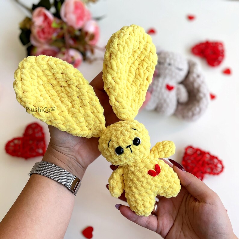May include: A yellow crocheted bunny with a red heart on its chest. The bunny is being held by a hand wearing a silver watch. The bunny is surrounded by red hearts.